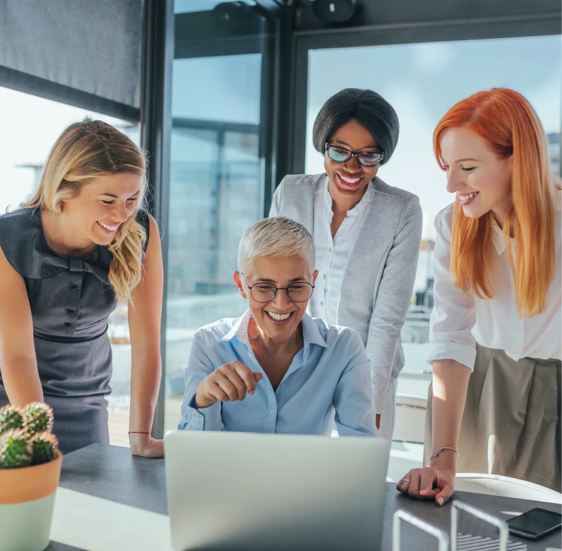 Four women in business attire smiling and gathered around a laptop in a modern office setting.