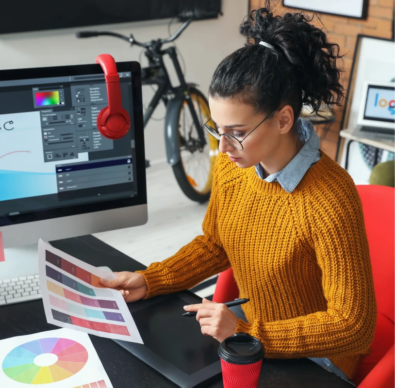 A person in a yellow sweater reviews color palettes at a desk with a computer, pen tablet, color charts, red headphones, and a bicycle in the background.