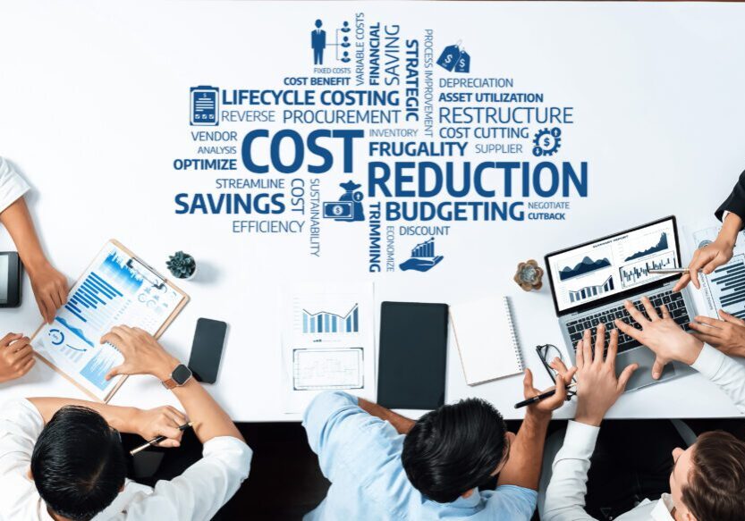 Top-down view of business people working at a table with laptops, tablets, charts, and a word cloud focused on cost reduction and financial planning.