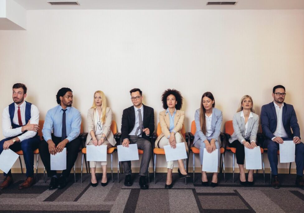 Seven professionally dressed individuals sit in a row of chairs, each holding papers, appearing to wait for a job interview.